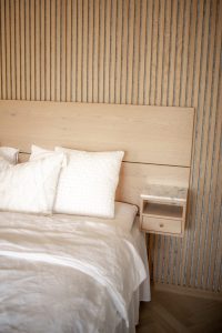 Imagem de quarto com soluções WoodUpp: painel ripado vertical em madeira clara, cabeceira lisa em carvalho claro e uma mesa de cabeceira suspensa WoodUpp com tampo de mármore e gaveta. Cama com roupa de cama branca e almofadas texturizadas sobre o colchão.