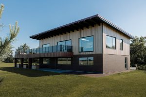 Casa moderna de dois andares com fachada superior revestida por ripas de madeira vertical WoodUpp, janelas amplas e varanda com guarda-corpo de vidro; base em alvenaria de tijolo e floreiras de madeira WoodUpp sob a varanda, em gramado aberto e céu azul.