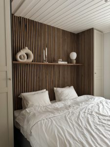 Quarto com painel ripado WoodUpp atrás da cama: painéis verticais de madeira escura com frestas pretas e prateleira estreita WoodUpp exibindo vasos cerâmicos abstratos, castiçais, um globo de vidro, livros e abajur de base bege. Roupa de cama e armários brancos completam o ambiente.