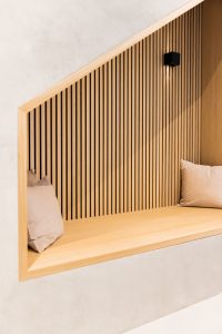 Recanto contemporâneo com painéis ripados WoodUpp em madeira clara formando parede inclinada, banco embutido WoodUpp do mesmo tom, duas almofadas bege e arandela de parede preta proporcionando luz dirigida. Acabamento minimalista, linhas verticais e moldura de madeira clara destacam o projeto.