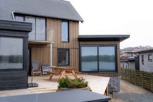 Fachada contemporânea WoodUpp com ripas verticais de madeira natural e caixilhos pretos, ampla varanda com deck de madeira WoodUpp, mesa piquenique em madeira e cadeiras de exterior. Vista para lago, floreira com plantas no primeiro plano e telhado cinza moderno.