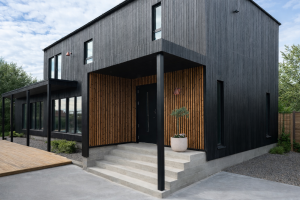 A imagem mostra uma casa moderna com fachada preta. A área da entrada coberta apresenta painéis ripados externos da WoodUpp em um tom de carvalho natural. Estas ripas verticais de madeira criam um contraste acolhedor contra o revestimento escuro da residência e as escadas de concreto.