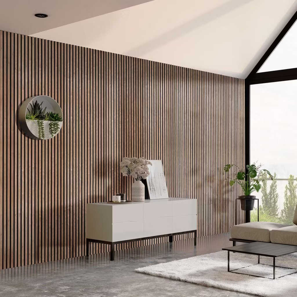 Interior minimalista com parede revestida pelos painéis ripados de madeira WoodUpp, com lâminas verticais e perfis escuros; aparador branco baixo, vaso com flores secas, quadro apoiado, sofá e plantas junto à janela, tapete claro e piso de cimento polido moderno.